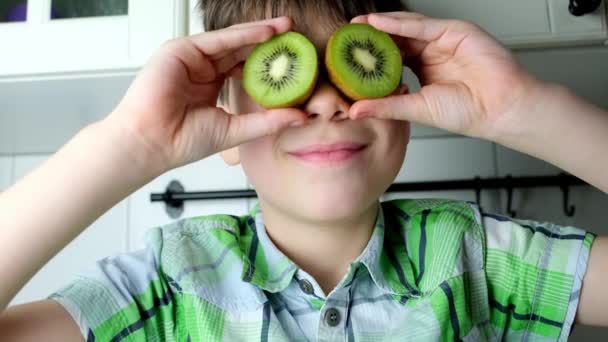 子供8 家のキッチンに座っている男の子 目の代わりにジューシーなカット緑のキウイ ヘッドクローズアップ 子供の食事のコンセプト 健康食品 — ストック動画