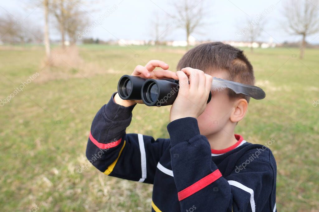 boy, guy 7-8 years old with a feather, stalker looks through black binoculars in the park, peeps, hunts down secrets, the concept of surveillance, observation of people and animals