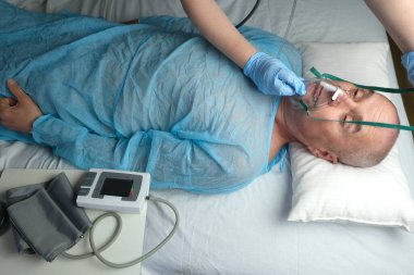 Mavi gömlekli yaşlı adam yalan söylüyor, doktor maske takıyor, tıbbi bakım konsepti, anestezi ve yoğun bakım, oksijen terapisi, koronavirüs tedavisi, COVID-19