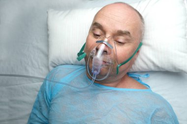elderly man patient in a blue shirt lies in a facial oxygen mask, the concept of medical care, anesthesiology and intensive care, oxygen therapy, resuscitation procedures clipart