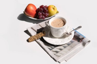 Beyaz bir fincanda kapuçino kahvesi ve beyaz arka planda çay tabağı, kurabiye, meyve, sabah gazetesi, sert ışık, kahve saati konsepti, güneşli gün, haber