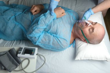 Mavi gömlekli yaşlı adam yalan söylüyor, doktor maske takıyor, tıbbi bakım konsepti, anestezi ve yoğun bakım, oksijen terapisi, koronavirüs tedavisi, COVID-19