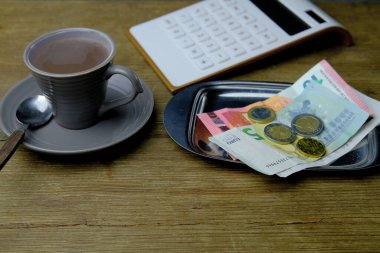 Tahta bir masada küçük gri bir fincan kahve, para için metal tepsi, euro banknotları ve bozuk paralar, hesap makinesi, restoran faturası, bahşiş parası konsepti, ev ekonomisi, gıda tasarrufu.