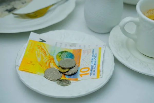 close-up of white plate for money, Swiss francs banknotes and coins, Restaurant bill, cup of coffee, delicate pink flowers, dishes with food, the concept of tip money, change of the waiter