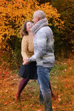 Yaşlı adam ve kadın, sonbahar parkında ağaçların arka planında dans ederken eğleniyorlar. Sağlıklı yaşam tarzı, iyi ruh hali, her yaşta mutluluk.