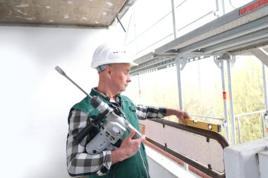 Koruyucu beyaz kasklı ve işçi üniformalı yaşlı inşaatçı inşaat ve onarım için inşaat alanında duruyor. İnşaat mühendisliği konsepti.