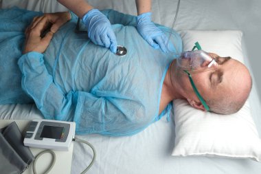 Mavi gömlekli yaşlı adam yalan söylüyor, doktor maske takıyor, tıbbi bakım konsepti, anestezi ve yoğun bakım, oksijen terapisi, koronavirüs tedavisi, COVID-19