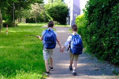 İki erkek çocuk, ilkokul öğrencileri, mavi çantalı çocuklar el ele tutuşarak, geçmişteki evleri ve yeşil yaz ağaçlarını geçerek okula geri dönerler.