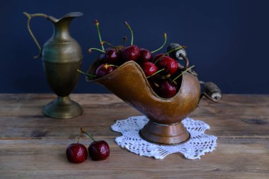 Koyu mavi arka planda küçük su damlalarında olgun kiraz, klasik bakır kül kapları, sürahiler mevsimlik meyve konsepti, sıcak yaz tazeliği, susuzluk giderme, yaz mevsimi