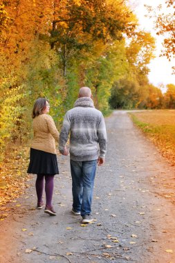Gri kazaklı orta yaşlı bir adam ve sarı stantlı bir kadın, bir sonbahar parkında, ağaçların arka planına sarılıyorlar. Sağlıklı yaşam tarzı, aşk, mutluluk kavramı her yaştan.