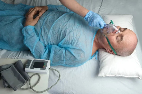 elderly man patient in blue shirt lies, the doctor puts on facial oxygen mask, concept of medical care, anesthesiology and intensive care, oxygen therapy, treatment of coronavirus, COVID-19