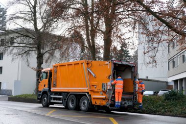 LUCERNE, SWITZERLAND - January 2021: orange cargo garbage truck driving through city streets, collecting garbage from bins, waste recycling concept, traffic safety regulation concept on streets clipart