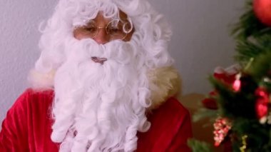 Geleneksel kırmızı sakallı Noel Baba süslemeli bir Noel ağacının yanında oturuyor, hediyelerle dolu kutular, mutlu yıllar diliyor, tebrik, mutlu noeller, mutlu noeller