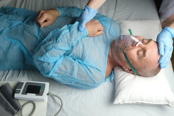 elderly man patient in blue shirt lies, the doctor puts on facial oxygen mask, concept of medical care, anesthesiology and intensive care, oxygen therapy, treatment of coronavirus, COVID-19