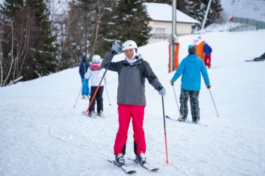 Beyaz kasklı ve kışlıklı gülümseyen adam karlı yamaçta duruyor. Aktif son sınıf öğrencisi, açık hava kış sporlarında kayak, sağlık, enerji ve neşeyi seviyor. Kayak sopaları tutuyor ve arka planda kayakçılar var.