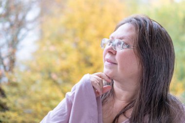 Sonbahar parkında düşünceli, nötr bir ifadeye sahip olgun bir kadın yüzü nötr duygulara sahip, gözlerinin etrafında kırışıklıklar, yumuşak doğal ışık, sakin ruh hali, mevsimsel portre konsepti