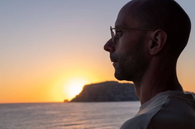 Gözlüklü bir adam Akdeniz 'de altın gün batımını seyrediyor, ılık gökyüzüne karşı siluet, sakin deniz manzarası, tefekkür, huzur ve sahil sükunetini çağrıştırıyor, huzur dolu deniz manzarası