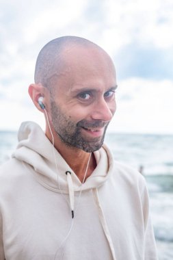 Mutlu genç adam deniz kenarında müzik dinlerken, gülerken ve sahilde göz kırparken, kulaklıkla müzik dinlerken, mizahi iyimser portre, neşeli ve rahat tatil havasında