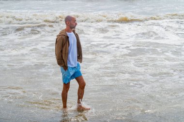 Genç adam köpüklü okyanus dalgalarında duruyor, erkek sörf yaparken huzuru buluyor, ritim dalgalarının endişeleri yıkamasına izin veriyor, şort ve ceket giyiyor, dinginliği somutlaştırıyor, doğayla bağlantı kuruyor, sahil kaçışı yapıyor.