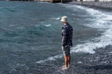 İçgözlemci adam dizlerine kadar turkuaz sulara batmış, kareli gömlek ve soluk kova şapkalı sahil gezgini, doğanın ritim duygusunu tecrübe ediyor.