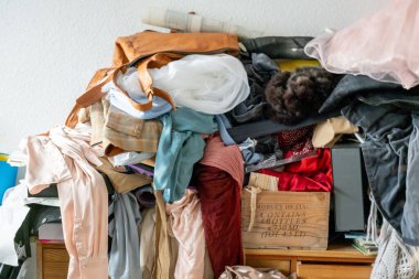 Karışık giysi ve kumaş yığını dağınık bir şekilde rafta ve ahşap sandıkta duruyor, dağınıklığı gösteriyor ve ev düzenine, dağınıklığına, depolama kaosuna, organizasyon ihtiyacına, gardıroba aşırı yüklenmeye ihtiyaç duyuyor.