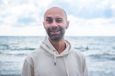 Mutlu genç adam deniz kenarında müzik dinlerken, gülerken ve sahilde göz kırparken, kulaklıkla müzik dinlerken, mizahi iyimser portre, neşeli ve rahat tatil havasında