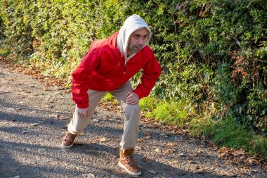 Kırmızı ceketli genç adam, bacak ve bacak, park yolunda çömelmiş, koşma öncesi dinamik esneme, soğuk hava, disiplin ve küçük günlük zaferler, yeşil çalılarla çevrili, tek başına eğitim yapıyor.