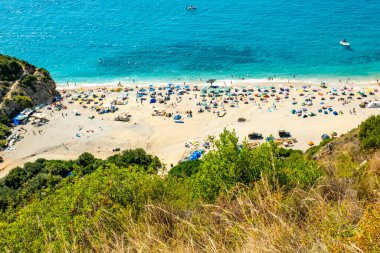 Turkuaz kıyı şeridi boyunca şemsiye altında insanlarla dolu canlı bir plaj sahnesi, turistler güneşli ve denizin keyfini çıkarıyor, Arnavutluk tatili, doğal güzellik insan aktiviteleri, trend Balkanlar turizmi ve Arnavutluk 'la tanışıyor