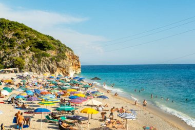 Turkuaz kıyı şeridi boyunca şemsiyelerin altında insanlarla dolu canlı bir plaj sahnesi, turistler güneş ve denizin keyfini çıkarıyor, Arnavutluk tatili, Doğal güzellik insan aktiviteleriyle tanışıyor, Vlore, Arnavutluk - 7 Eylül 2025