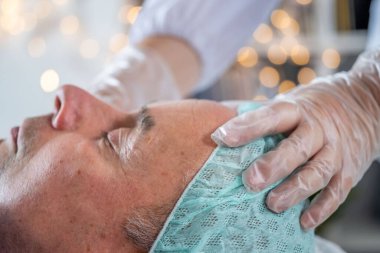 Terapistin elleri yaşlı bir adamın yüzüne masaj yapıyor, tirajı ve deri esnekliğini arttırıyor, uzman adam profesyonel olarak rahatlatıcı yüz masajı yaptırıyor güzellik salonu, kaplıca ve sağlık konsepti alıyor.