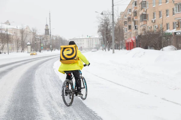 13.02.2021, Russia, Moscow. A food deliveryman on a bicycle crosses the road in winter. Snowfall and snowdrifts — Stock Photo, Image