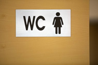 Üzerinde siyah harflerle WC çeliği olan sarı ahşap bir duvar ve kadın insan ikonu. Tuvalet işaretçisi.