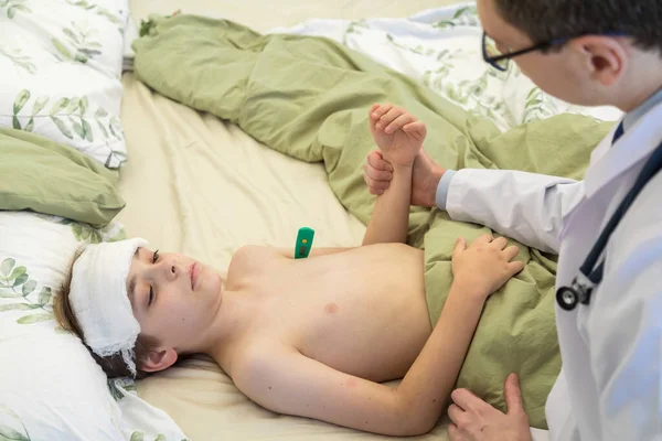 O médico examina o paciente com a temperatura, resfriando a bandagem molhada na cabeça dos adolescentes. Um menino jaz na cama — Fotografia de Stock