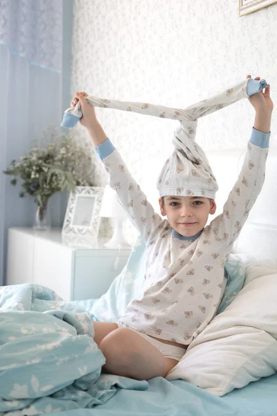 Niño caucásico en la mañana en la cama juega. El niño se pone pantalones de pijama en la cabeza y se divierte. —  Fotos de Stock