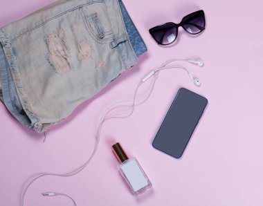 Cep telefonu, güneş gözlüğü, beyaz kulaklık, bir kız için hippi kot şortu açık pembe arka planda cam bir şişede parfüm. Yukarıdan görüntüle