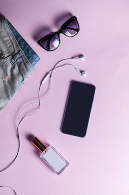 Cep telefonu, güneş gözlüğü, beyaz kulaklık, bir kız için hippi kot şortu açık pembe arka planda cam bir şişede parfüm. Yukarıdan görüntüle