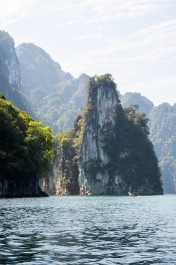 Arka planda sisli orman dağları olan su seviyesinde yakalanan küçük uzun kuyruklu bir tekne geçerken, durgun turkuaz sudan yükselen yüksek kireçtaşı karst adası. Khao Sok