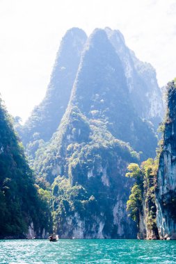 Uzun kuyruklu tekne gölde hız yapar. Ormanla kaplı yüksek uçurumlar ve turkuaz suyla çerçevelenmiş, gün ışığında alçak ve merkezli bir perspektiften yakalanmış..
