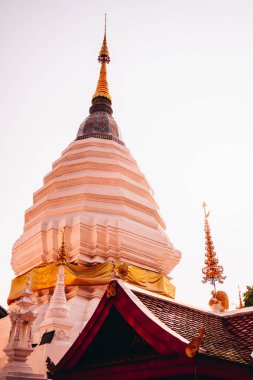 Chiang Mai. Geleneksel bir tapınak çatısının üzerinde yükselen altın kuleye sahip beyaz bir Budist 'in düşük açılı görüntüsü, sıcak ışıkla vurgulanmış ve Tayland mimarisinin detaylarıyla süslenmiş..