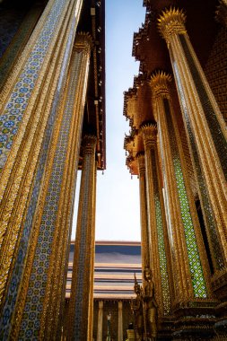 Wat Phra Kaew tapınağı. Bangkok 'ta. Karmaşık mozaik desenler ve oyulmuş detaylarla süslü altın tapınak sütunlarının alçak açılı görüntüsü gökyüzüne doğru yükseliyor.