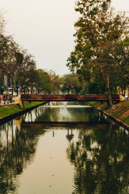 Sakin bir şehir kanalı ağaçları ve çiçeklerle süslenmiş küçük tahta bir köprüyü yansıtır ve yumuşak akşam ışığında yeşilliğin çerçevelediği huzurlu bir şehir vahası yaratır. Chiang Mai, Tayland