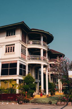 Kıvrımlı balkonları, beyaz sütunları ve çiçekli bitkileri olan büyüleyici bir koloni tarzı bina berrak mavi bir gökyüzünün altında duruyor, klasik mimariyle tropikal çevreyi harmanlıyor..