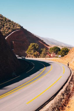 Sarmal asfalt dağ yolu, parlak mavi gökyüzü altında sarp kayalık bir uçurum etrafında kıvrımlar, düşük perspektiften çekilir, yalnızlığı, seyahati ve dramatik doğal manzarayı vurgular..
