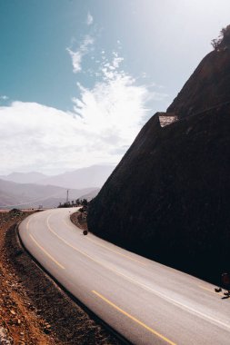 Sarmal asfalt dağ yolu, parlak mavi gökyüzü altında sarp kayalık bir uçurum etrafında kıvrımlar, düşük perspektiften çekilir, yalnızlığı, seyahati ve dramatik doğal manzarayı vurgular..