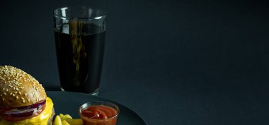 Patates kızartmalı burger ve siyah arka planda bir bardak kola yanında ketçap.