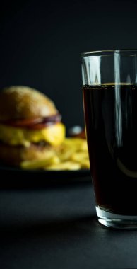 Bulanık hamburgerin yanında bir bardak kola ve siyah arka planda patates kızartması.