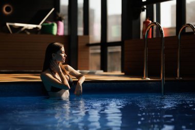 Esmer kadın, spa merkezindeki havuz başında gün ışığında duruyor.