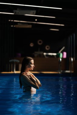 Side view of brunette woman standing in sunlight in blue swimming pool