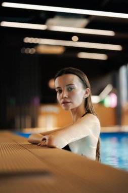 Islak saçlı bir kadın, spa merkezindeki havuz kenarında kameraya bakıyor.