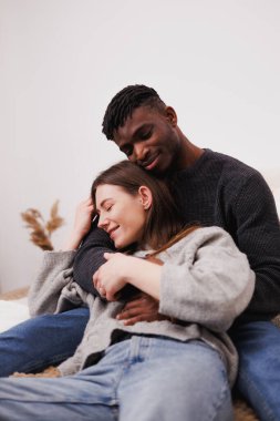 Gülümseyen ve rahat çok ırklı bir çift evde yatakta sarılıyor.
